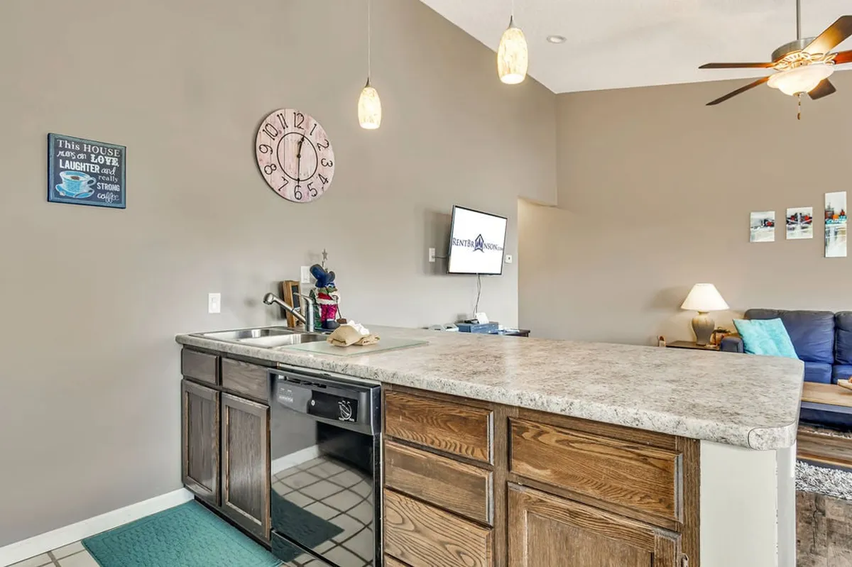 A bright, open-plan living and kitchen space with a kitchen island, pendant lighting, wall clock, television, and a comfortable sofa, offering plenty of room for socializing, watching TV, or hosting game nights after a busy day out.