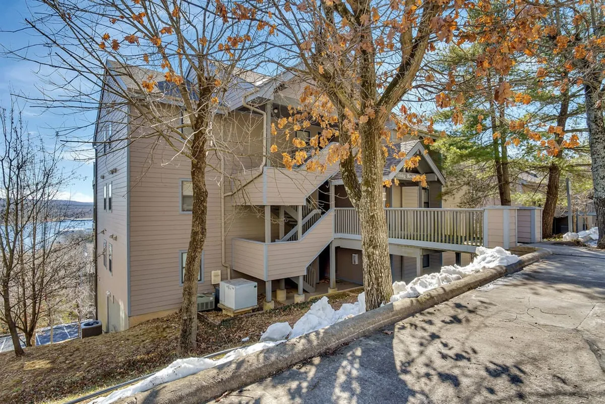 An exterior view of the building nestled among trees, with a driveway leading up and a porch that suggests relaxation and enjoying the outdoors. The tranquil setting hints at natural beauty and proximity to lakeside activities.
