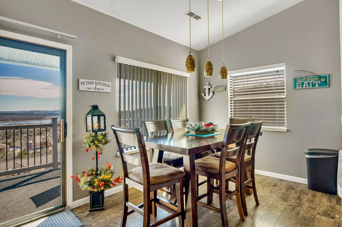 A dining table is set near a sliding glass door that leads out to a balcony or deck, framed by a decorative anchor and a bright floral arrangement. The scene radiates warmth, with the outdoor view and deck clearly visible, reinforcing the indoor-outdoor flow.