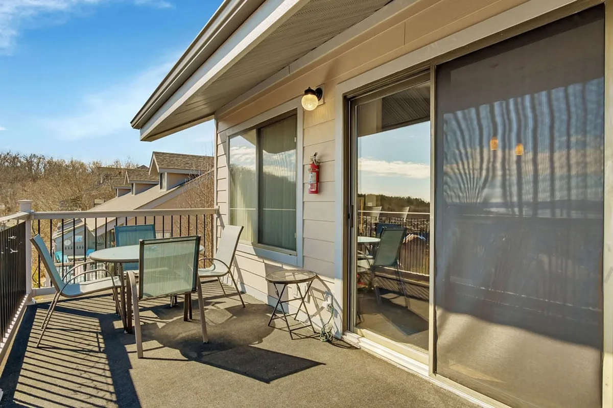 This exterior image features a balcony setup with a table and chairs overlooking a scenic outdoor view. Large windows behind the seating area connect the interior living space to the outdoors, capturing the tranquil atmosphere of lakeside living.