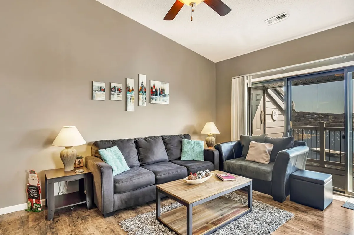 A cozy, inviting living room with a plush sofa and coffee table, accented by tasteful wall art. Sunlight floods the room through a sliding glass door, which opens to a deck boasting serene lake views, perfect for morning coffee or evening relaxation with loved ones.