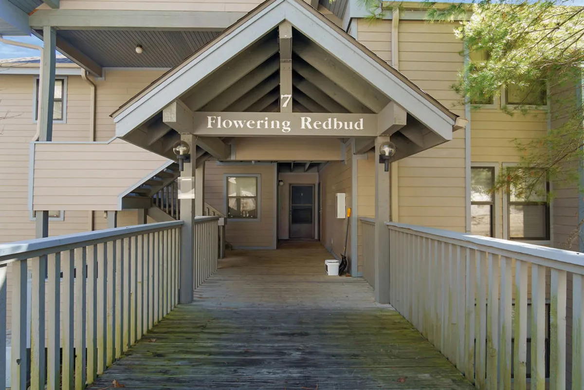This image presents the entrance sign 'Flowering Redbud' mounted on a wooden structure, with a wooden walkway and attractive overhang architecture. The immediate approach and friendly ambiance signal a welcoming arrival, inviting guests into the property and setting the tone for a getaway close to area attractions.