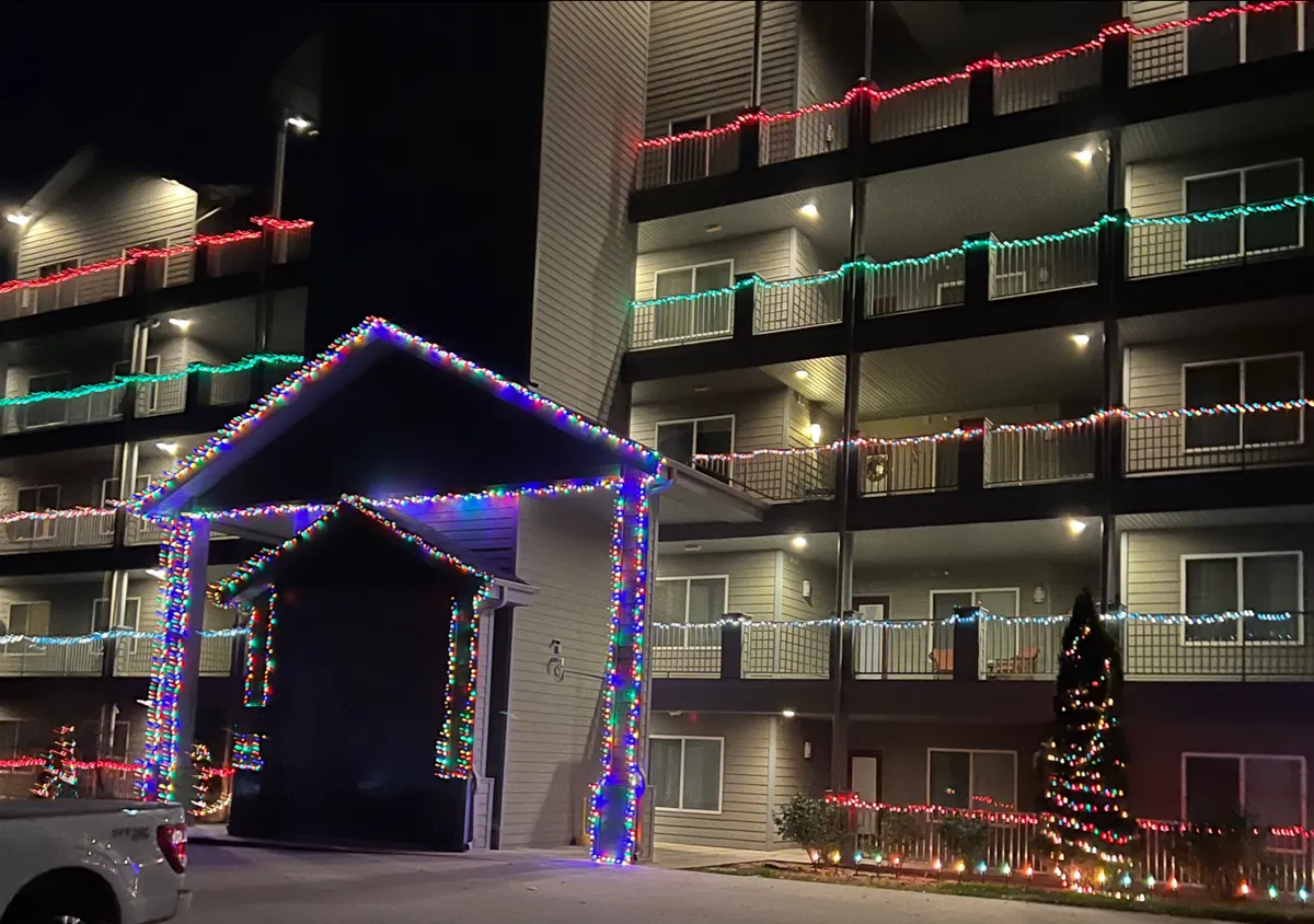 A warmly lit exterior entrance decorated for the holidays, featuring sparkling decorative lights, an inviting entrance canopy, and a prominently displayed Christmas tree. The setting exudes a sense of welcome, celebration, and togetherness.