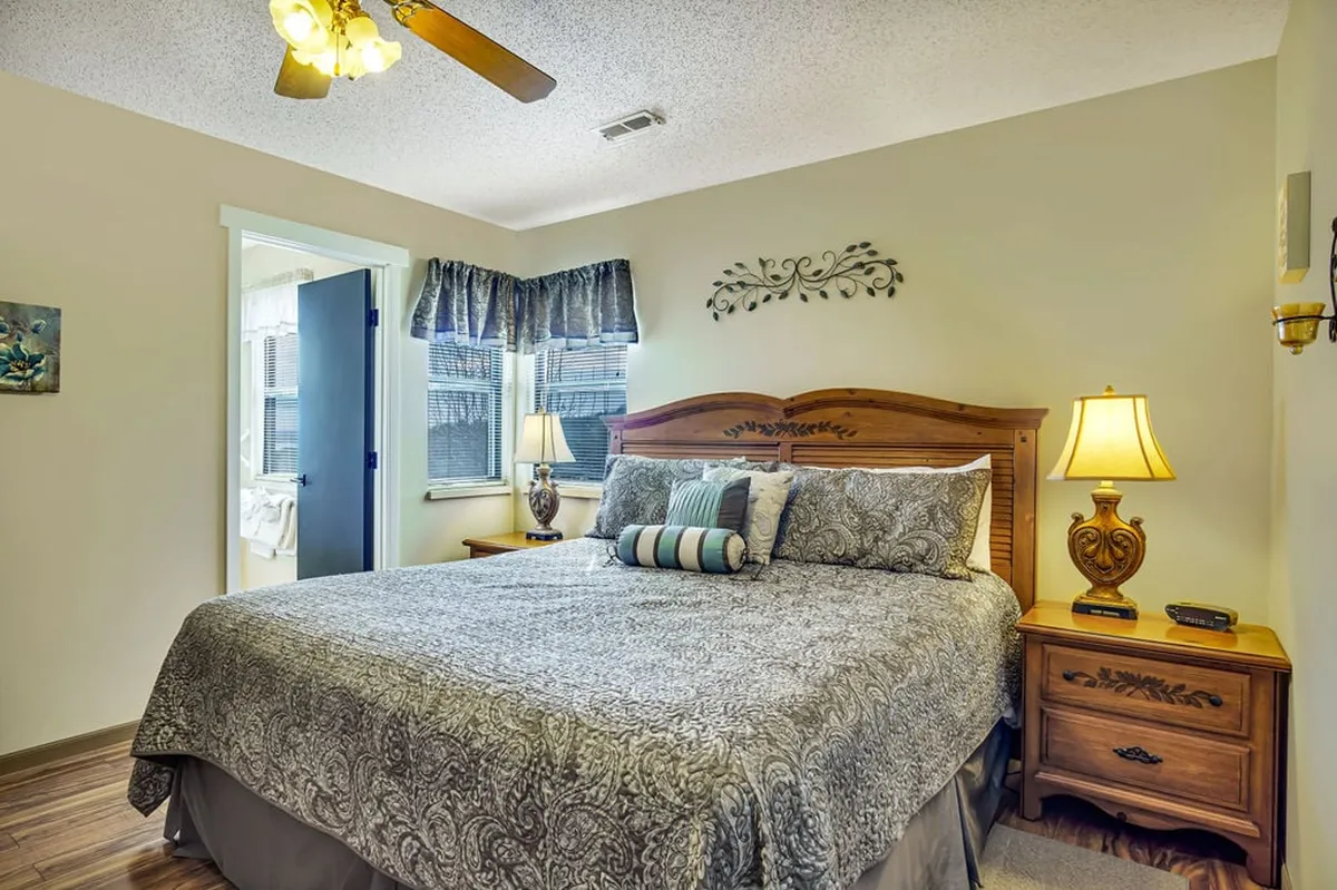 A serene and stylish master bedroom featuring a plush king-sized bed flanked by matching nightstands and cozy lamps, with a decorative wall piece above the headboard. Soft natural light filters through nearby windows, creating a tranquil atmosphere ideal for rest and relaxation.