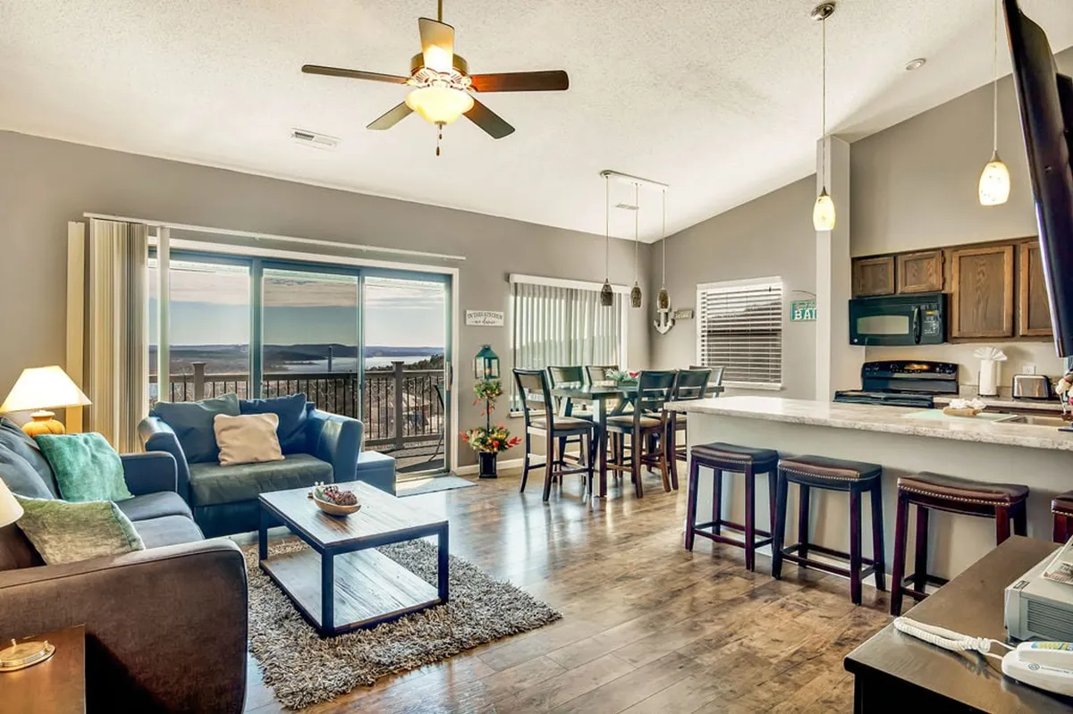 A spacious, light-filled area featuring a cozy seating area, a dining table with chairs, and a kitchen bar with stools. Expansive windows provide panoramic lake views, and the furnishings are modern and inviting. The space looks ideal for family gatherings, relaxation, and shared meals.